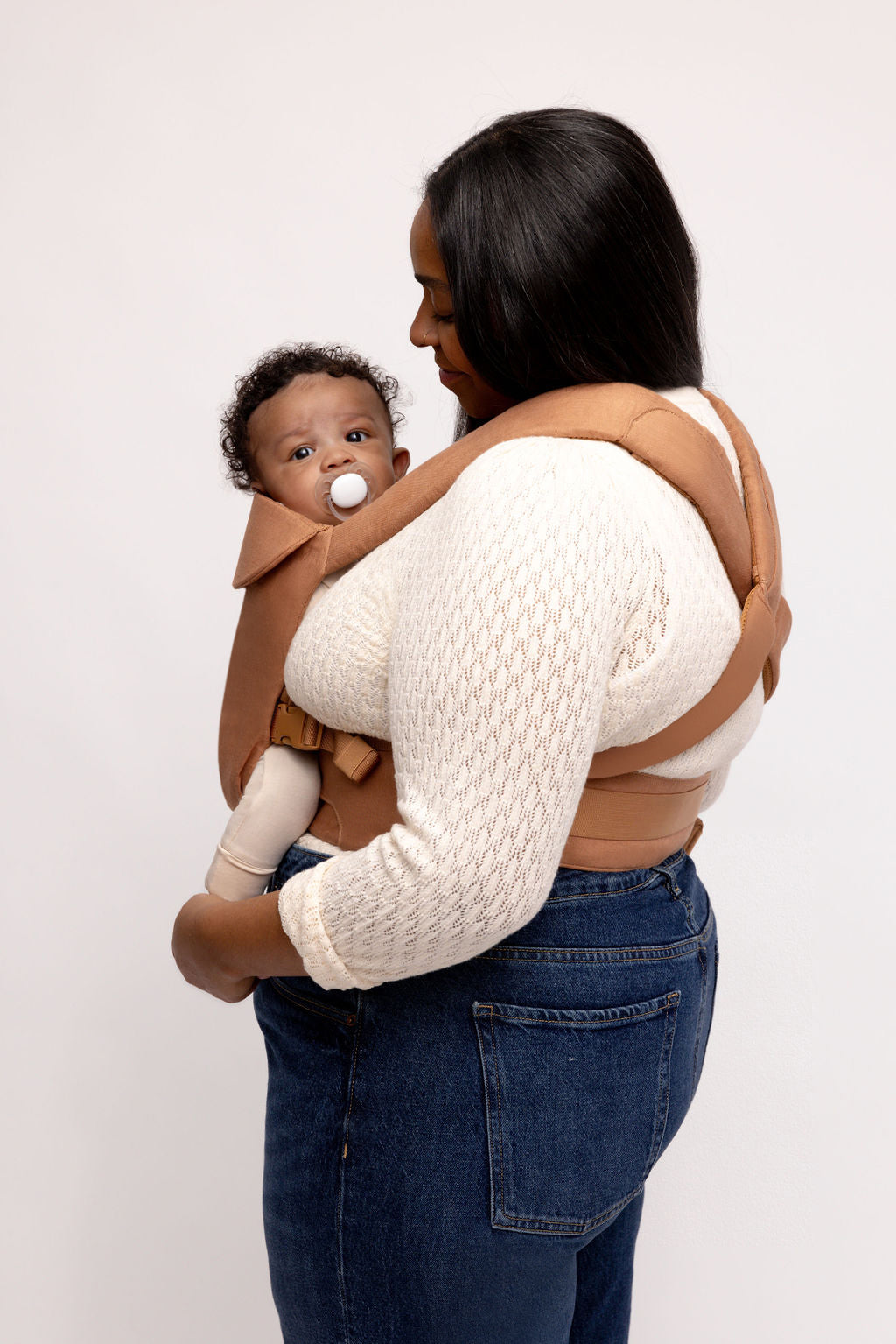 Mom holding baby in creamy caramel brown Aerial Carrier with matching straps and buckles, showcasing comfort and style.