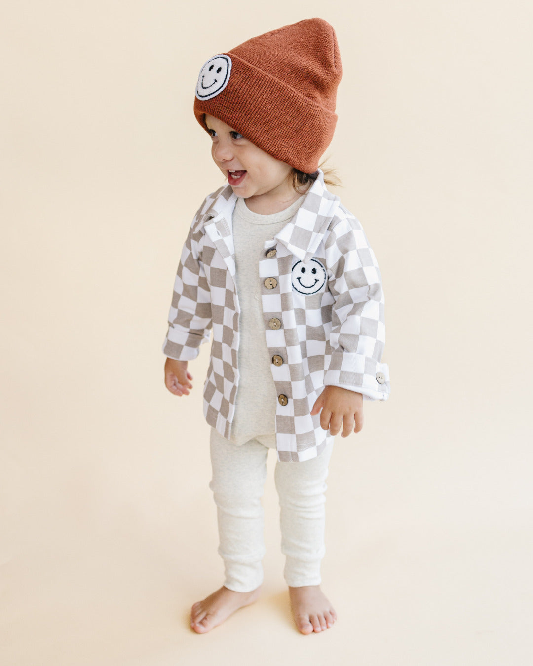 Toddler wearing a rust Smiley Beanie and a checkered shirt, spreading joy and warmth in cozy fashion.