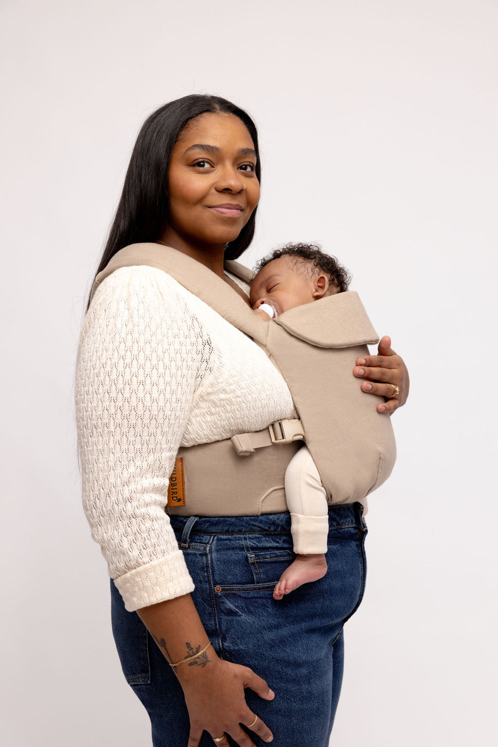 Woman wearing the Desert Lark Aerial Carrier in cool taupe grey, safely carrying her baby.