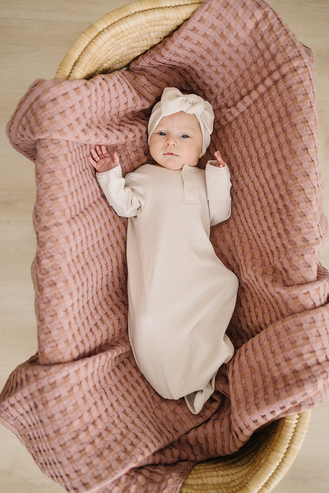 Sweet newborn in a vanilla organic baby knot gown, relaxing in a cozy basket with a textured blanket.