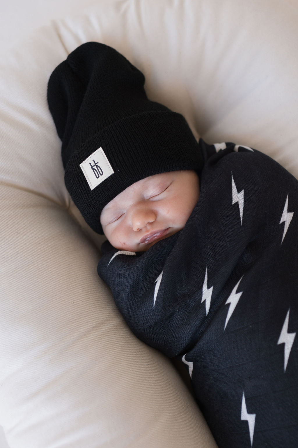 Baby peacefully sleeping in a Muslin Swaddle Midnight Lightning Bolt with a black beanie hat.