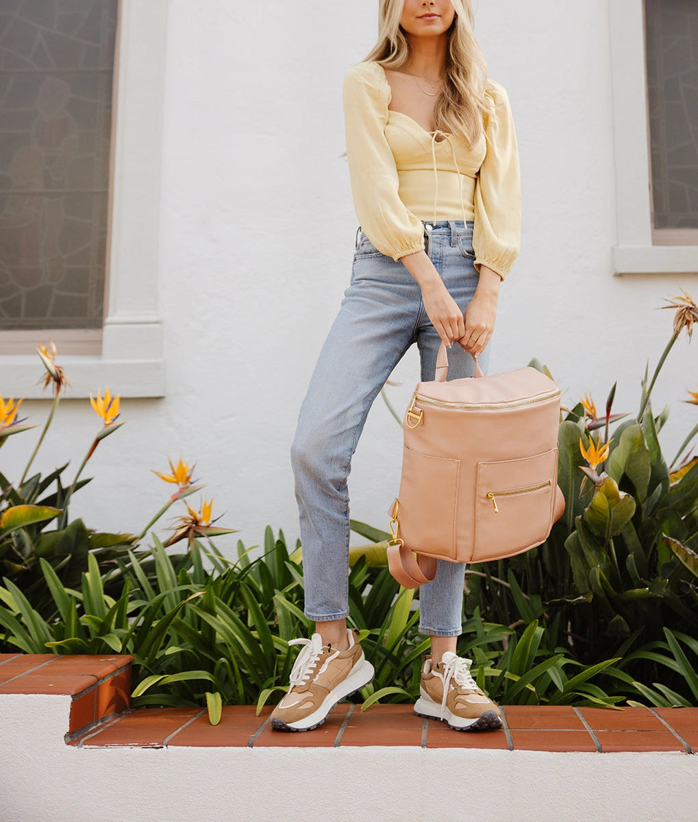 Model holding Original Diaper Bag Backpack in warm blush, styled with casual jeans and a yellow top, surrounded by greenery.