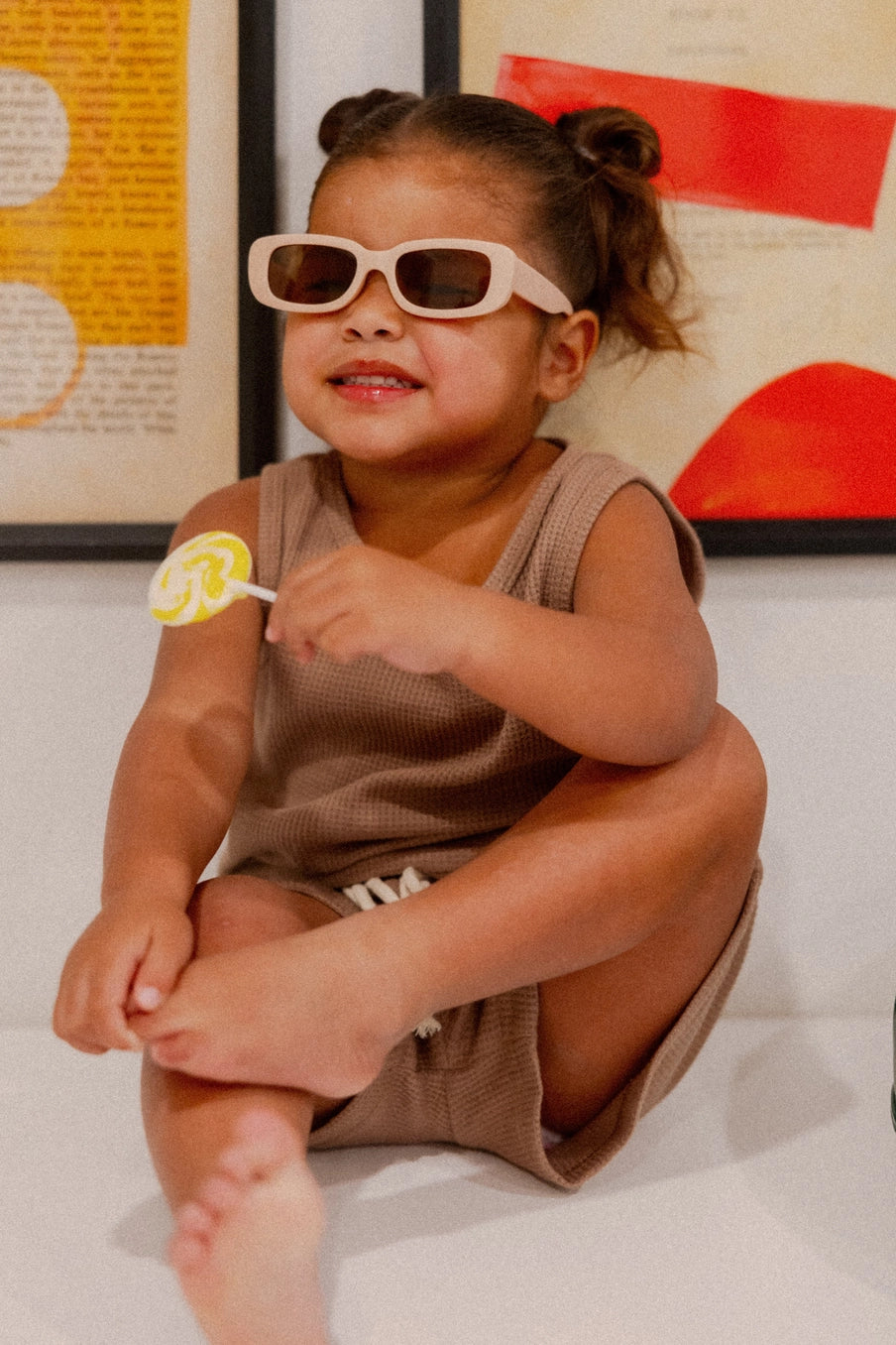 Smiling child in brown waffle-knit Madden Tank Set, wearing sunglasses, enjoying a lollipop indoors.