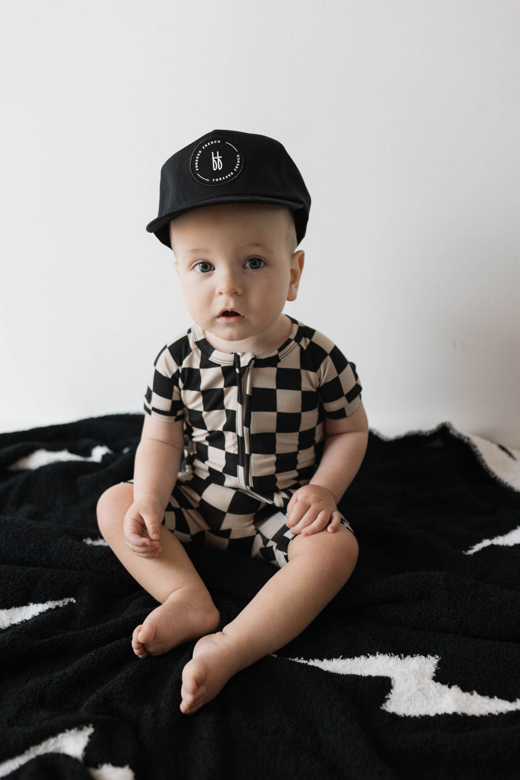 Baby wearing a black checkerboard bamboo zip shortie on a black and white blanket, showcasing a trendy summer outfit.