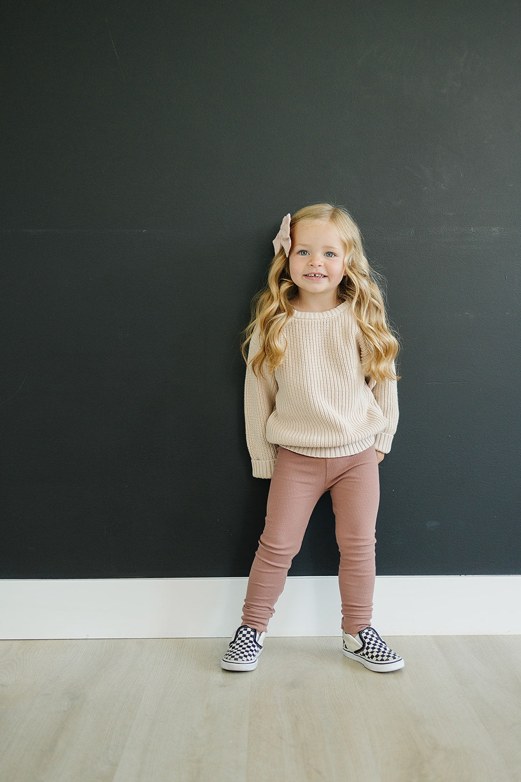 Cute girl wearing dusty rose organic baby leggings with a beige sweater and checkered shoes against a dark wall.