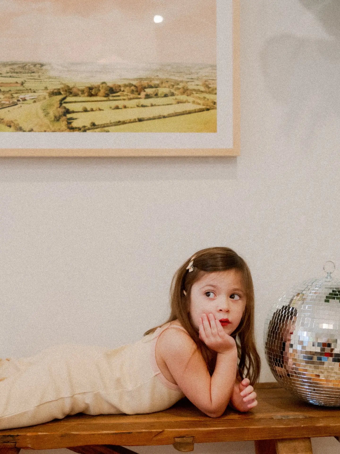 Little girl in a cream ribbed jumpsuit lounging on a wooden table next to a disco ball.
