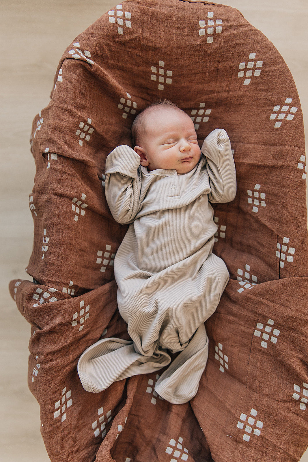 Newborn peacefully sleeping in an oatmeal organic baby knot gown on a patterned swaddle blanket.