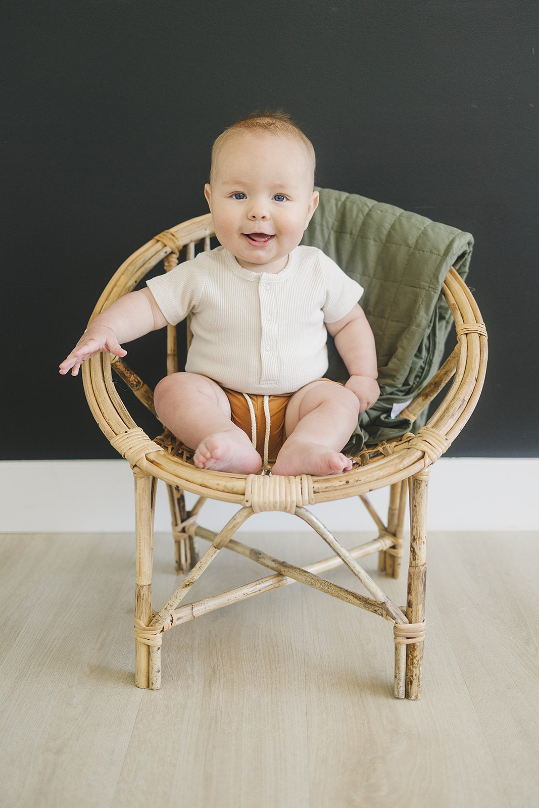 Baby in a vanilla organic baby bodysuit sitting in a rattan chair, showcasing a cute, minimalist outfit.