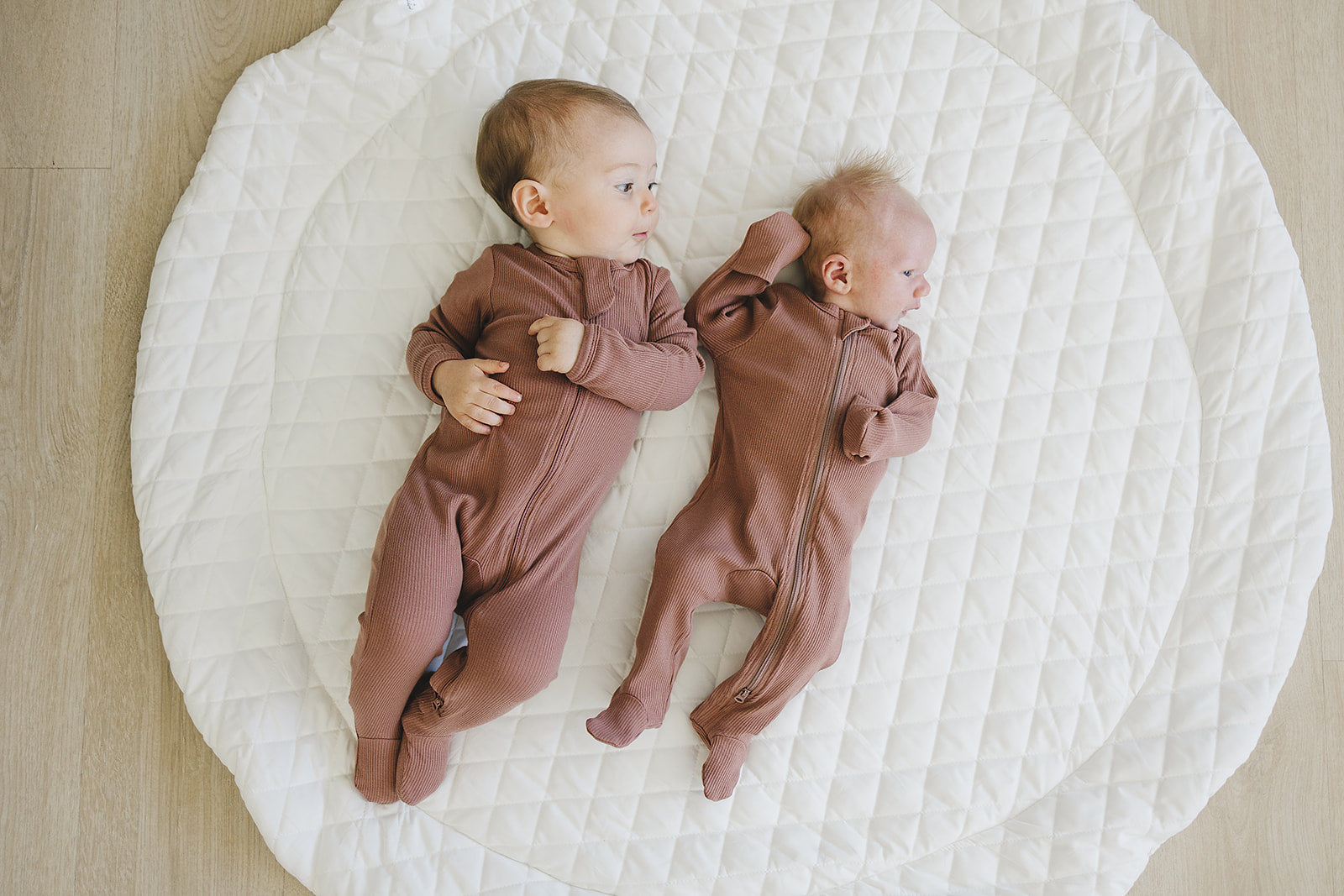 Two babies wearing dusty rose organic cotton zipper sleepers on a soft quilted blanket.