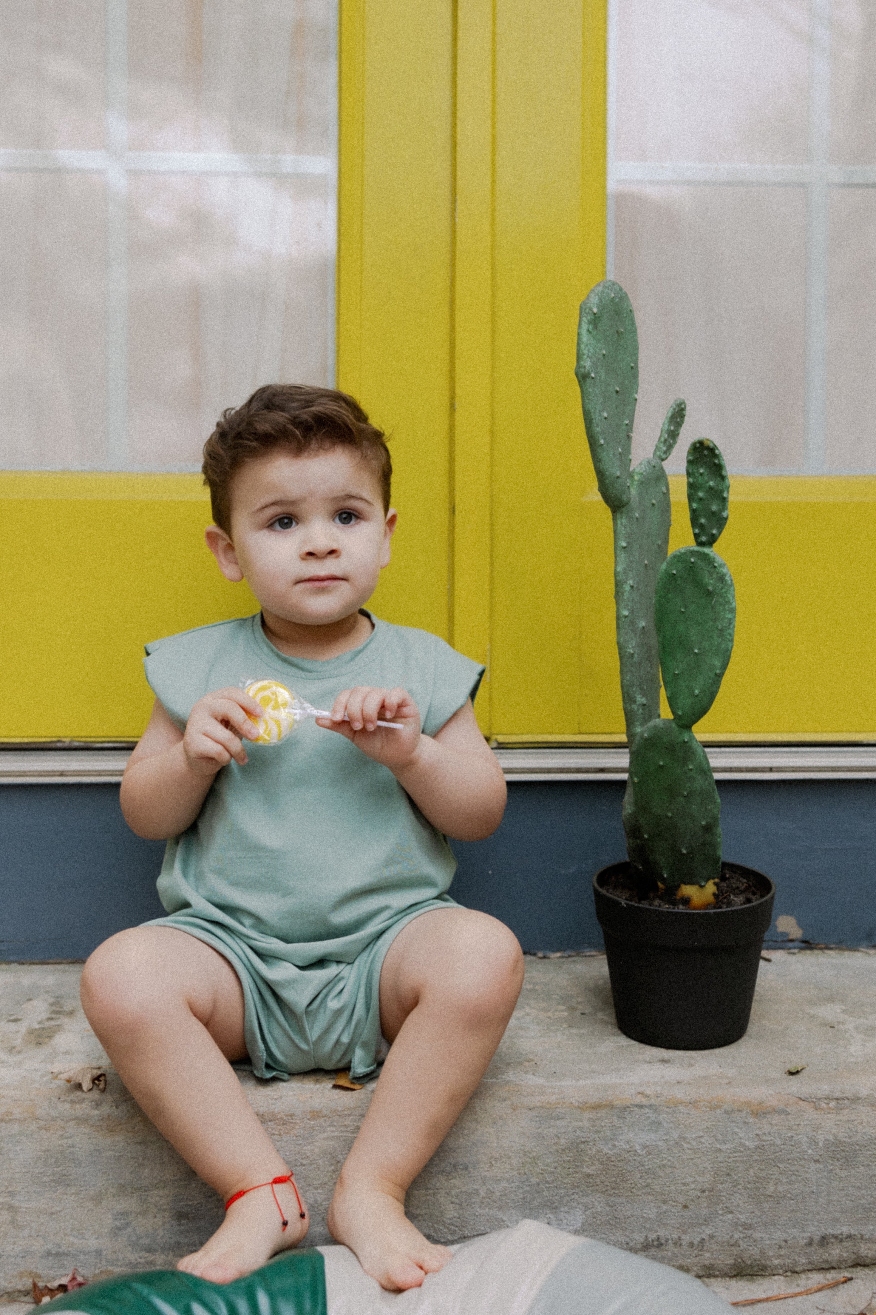 Toddler in teal Remy two-piece outfit sitting by a cactus near yellow doors, enjoying playtime in cozy cotton apparel.