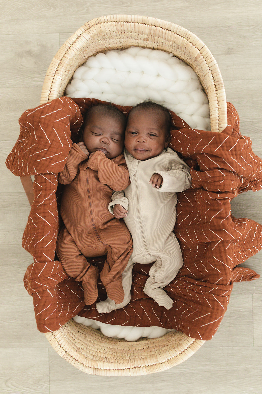 Two babies in organic cotton sleepers, one in rust and one in vanilla, nestled in a cozy basket.