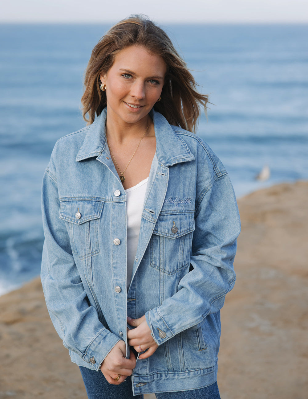 Woman wearing a What a Friend in Jesus denim jacket by the ocean, showcasing a faith-based fashion statement.