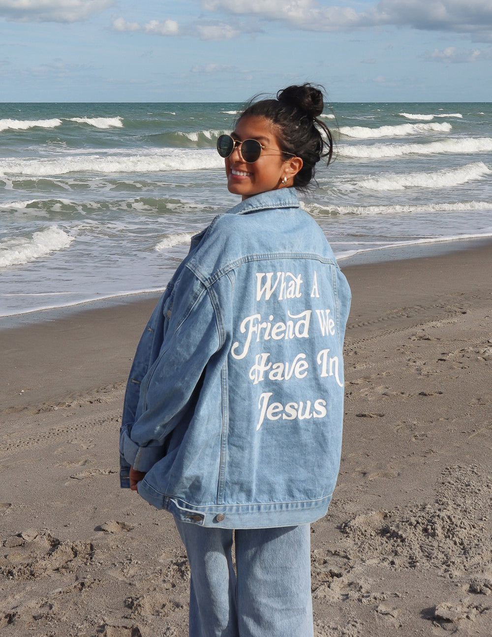 Woman wearing a 'What a Friend in Jesus' denim jacket on the beach, showcasing bold faith-based lettering.