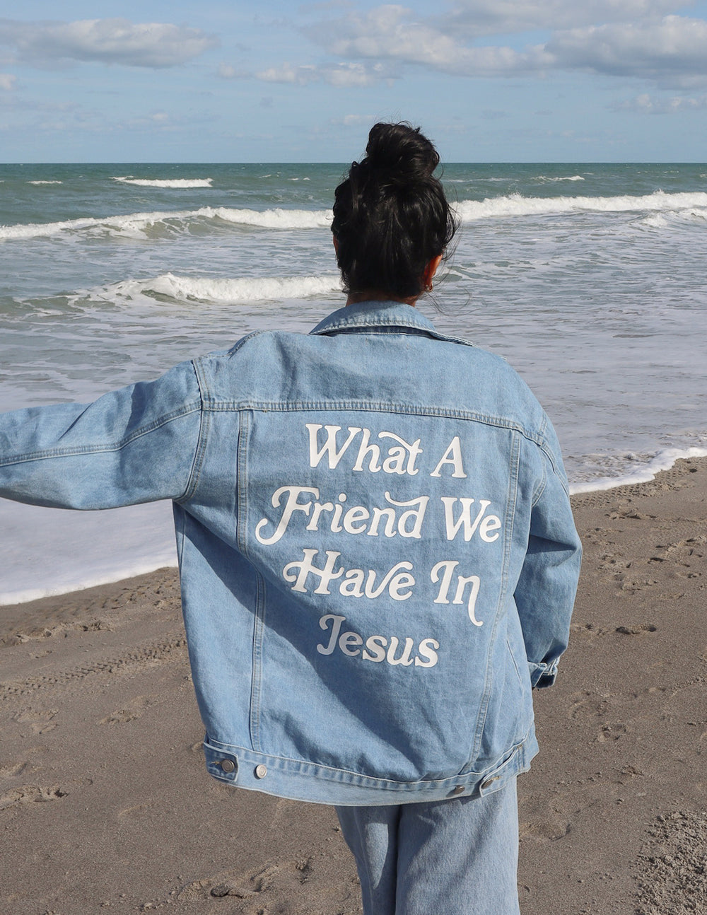 Woman wearing oversized denim jacket with 'What a Friend We Have In Jesus' text by the beach.