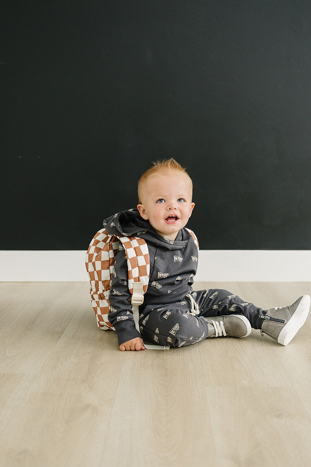 Toddler wearing a trendy outfit with a rust wavy checkered mini backpack, sitting on the floor happily.
