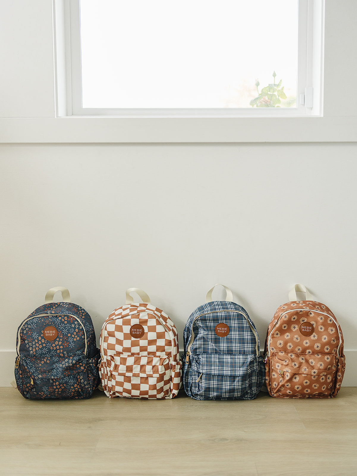Four stylish kids mini backpacks lined up against a wall, featuring various playful designs and colors.