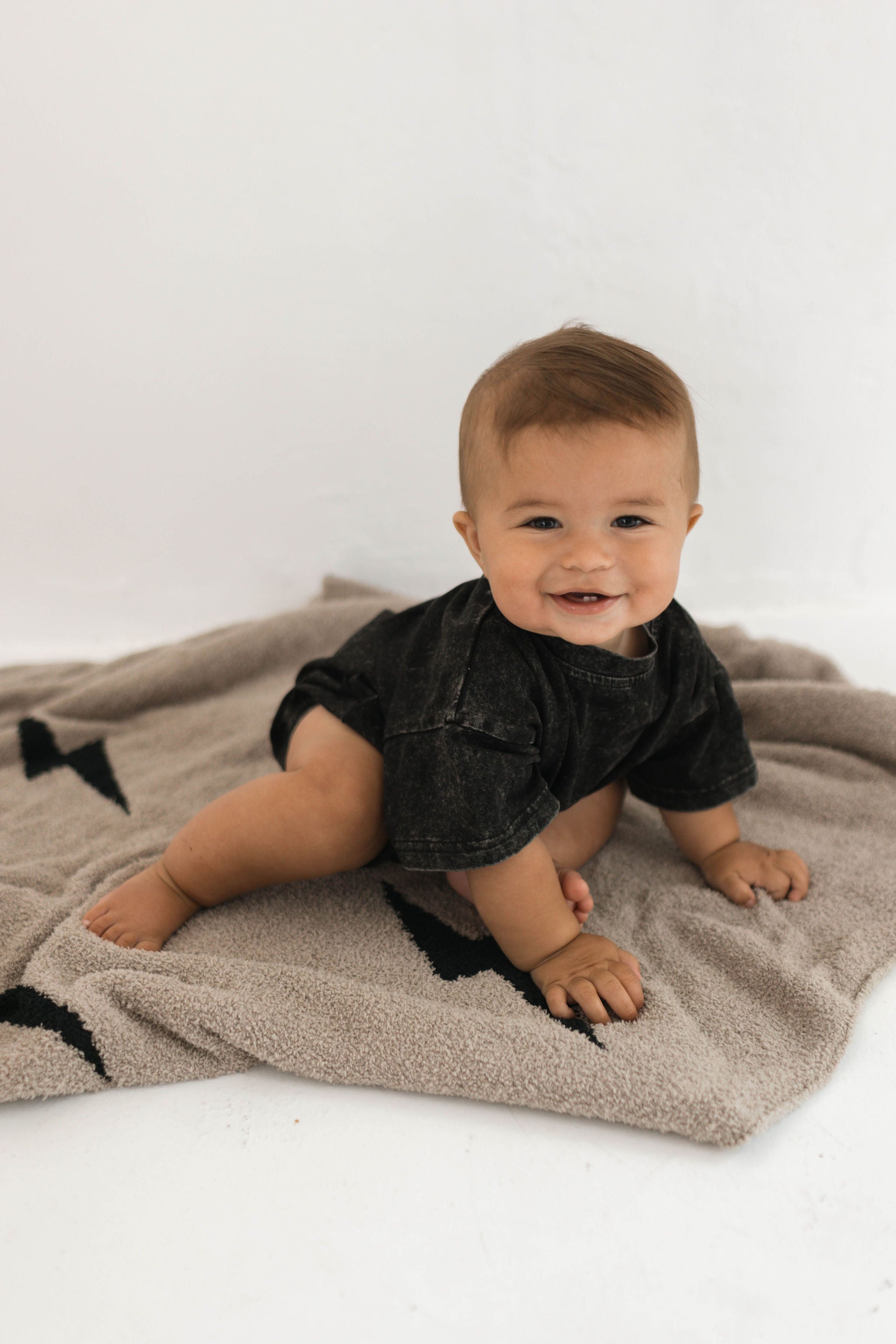 Baby smiling while sitting on a plush blanket with a lightning bolt design, wearing a black outfit.