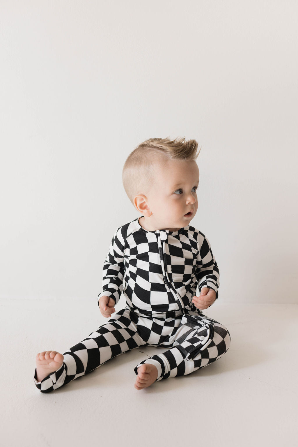 Black and white wavy checkerboard bamboo zip baby pajamas on a sitting child.