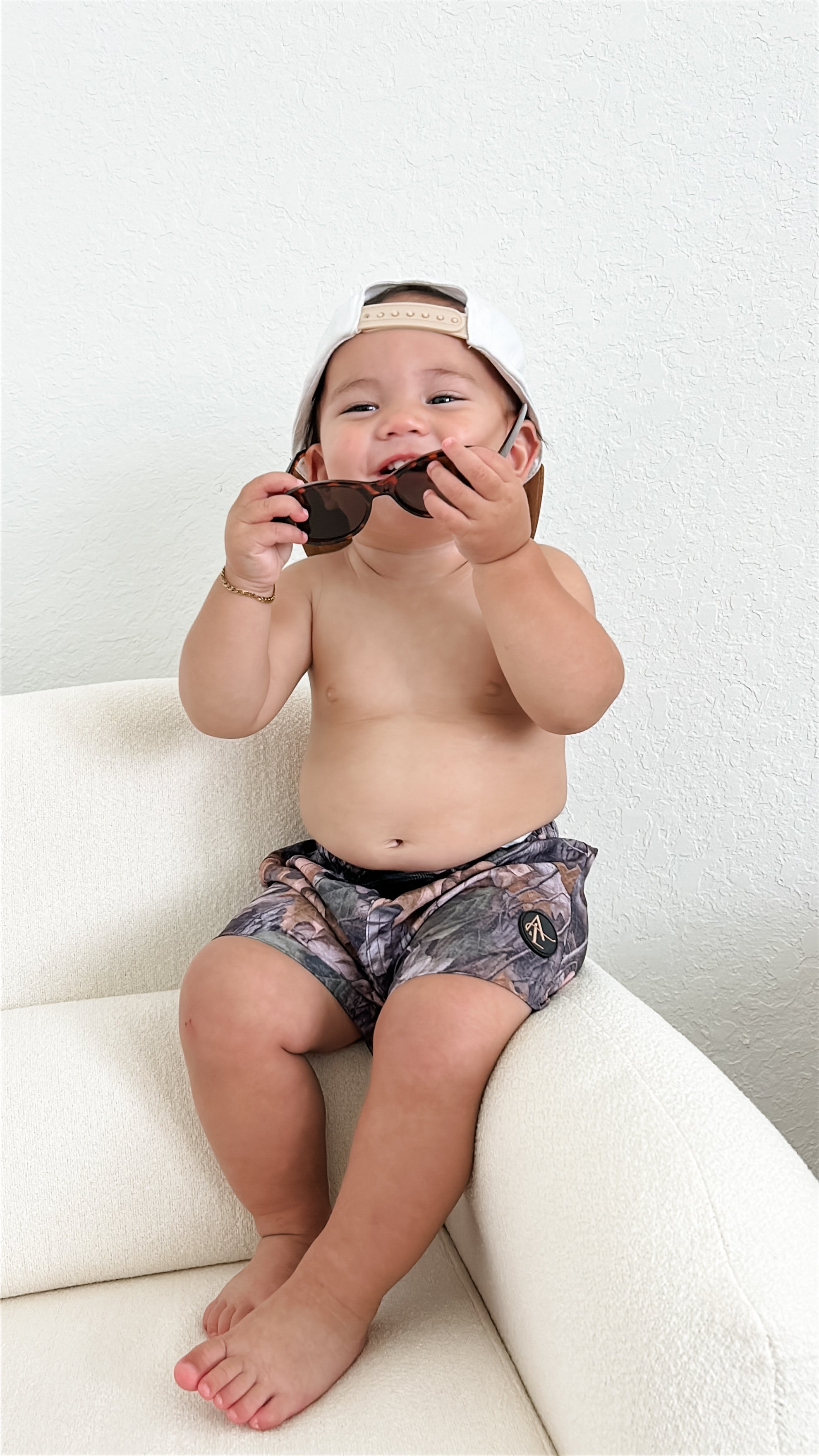 Toddler wearing camo swim shorts and sunglasses, sitting on a white couch, ready for summer water play.