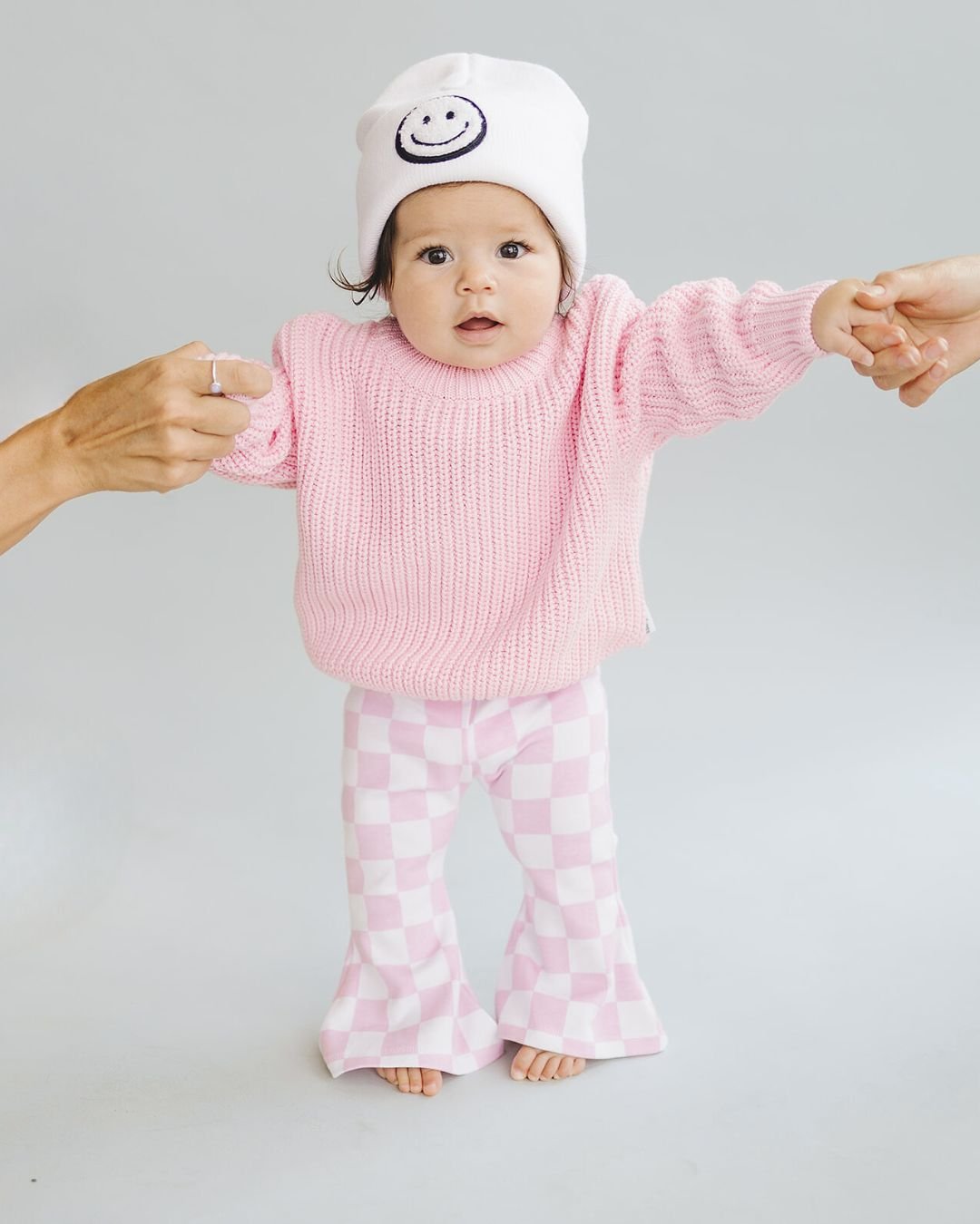 A baby wearing a pink chunky knit sweater and checkered pants, held by two adults, with a smiley face hat.