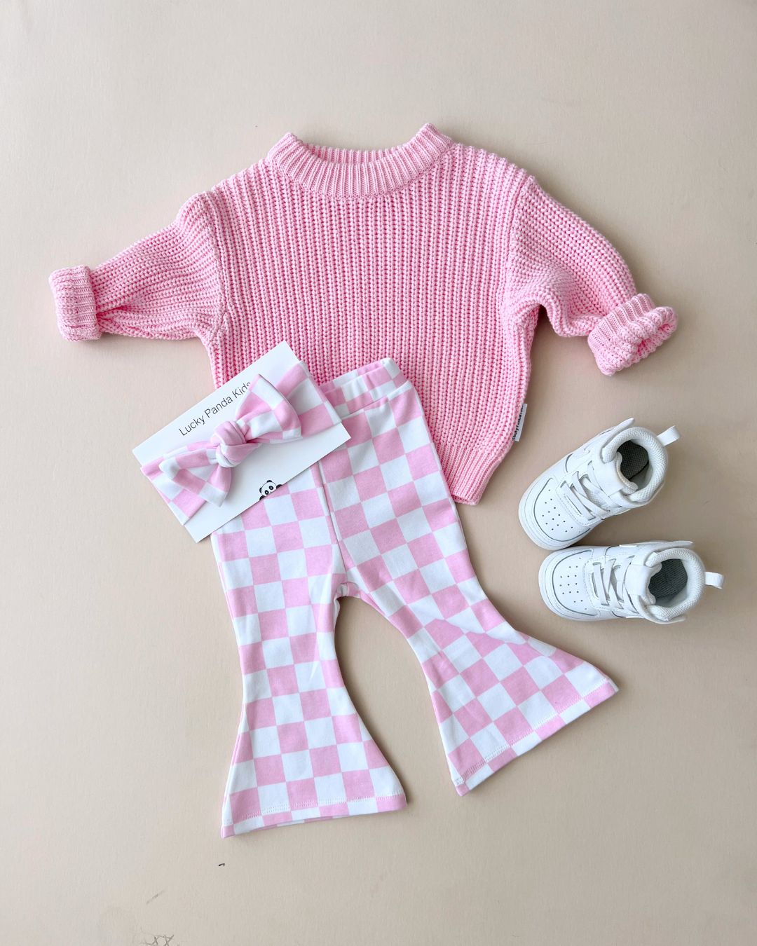 Chunky Knit Sweater in pink with checkered flared pants, a matching bow headband, and white sneakers on a light background.
