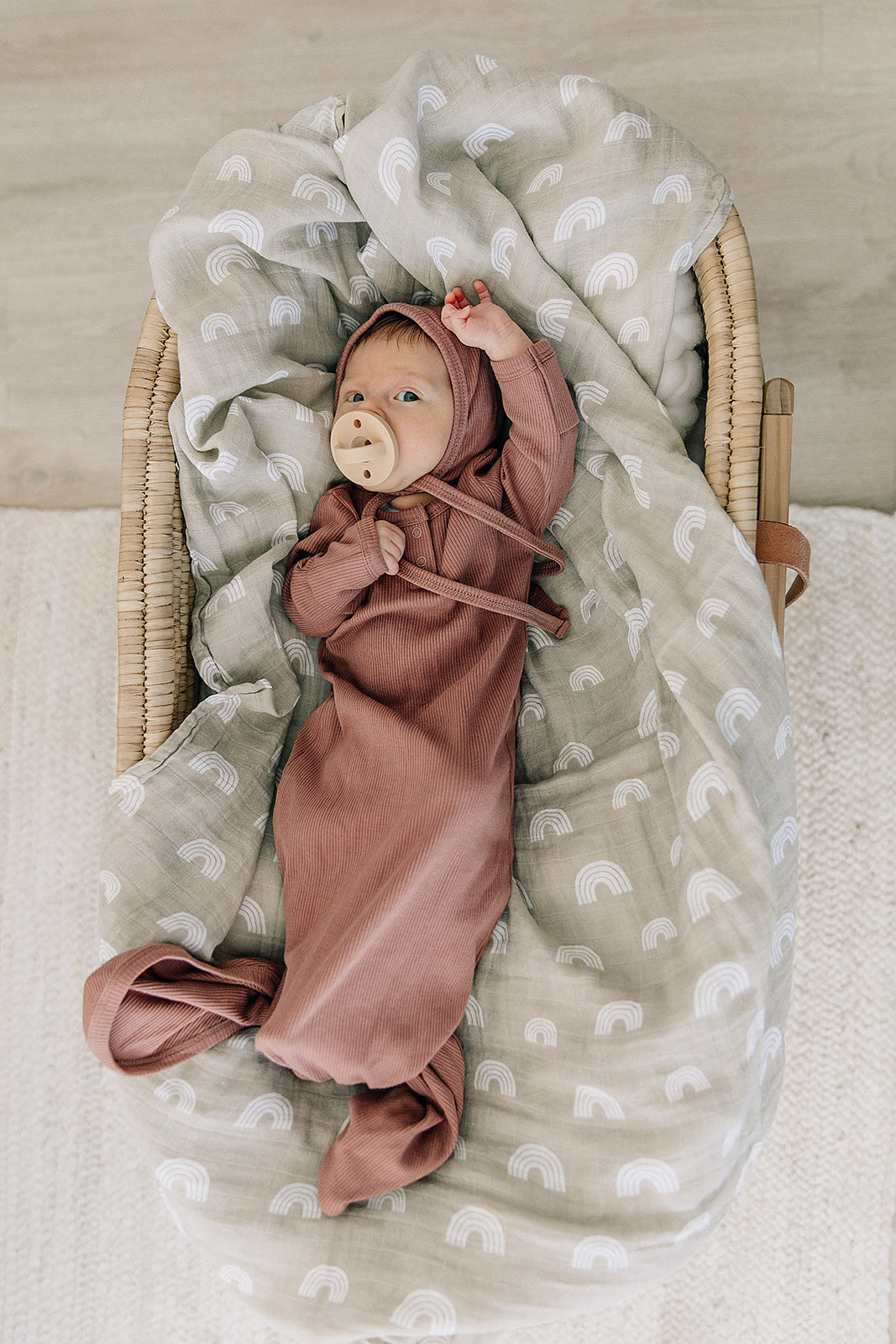 Newborn in a dusty rose organic baby knot gown, nestled in a cozy basket, showcasing adjustable knot bottom and fold-over mittens.