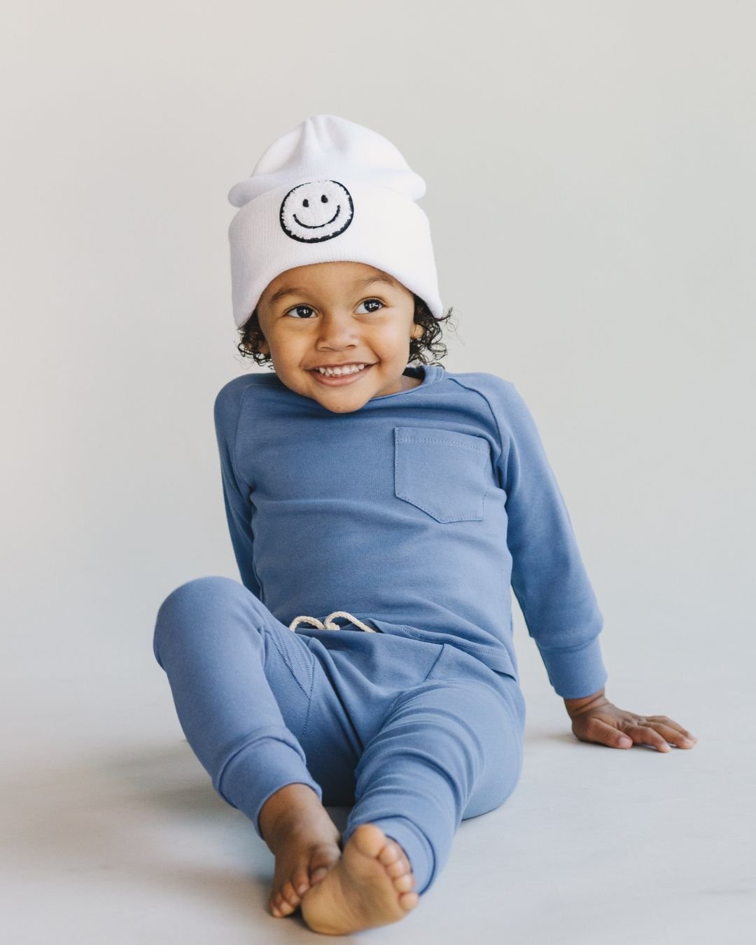 Toddler in lounge set ocean blue, smiling while wearing a white beanie with a smiley face.