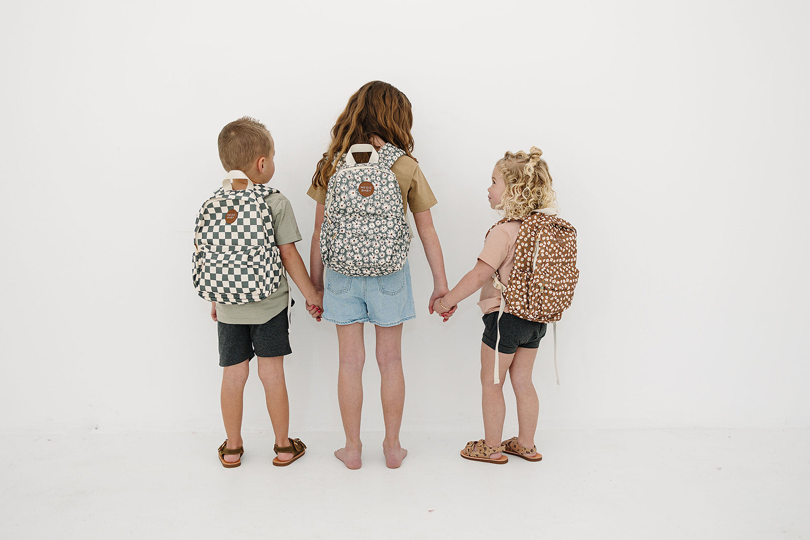 Three kids holding hands, each wearing stylish mini backpacks in playful patterns.