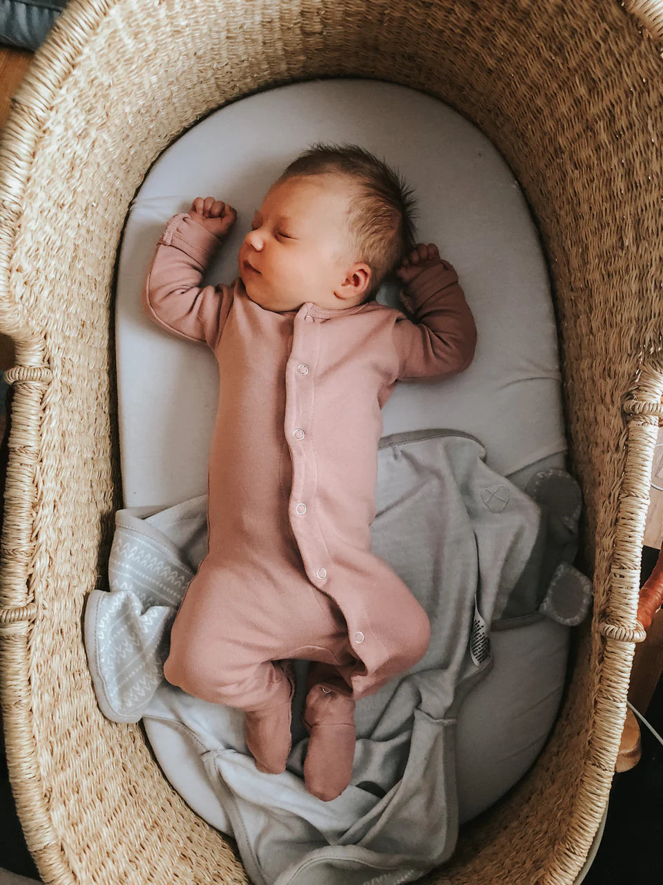 Baby wearing mauve organic snap footie while sleeping peacefully in a woven bassinet.