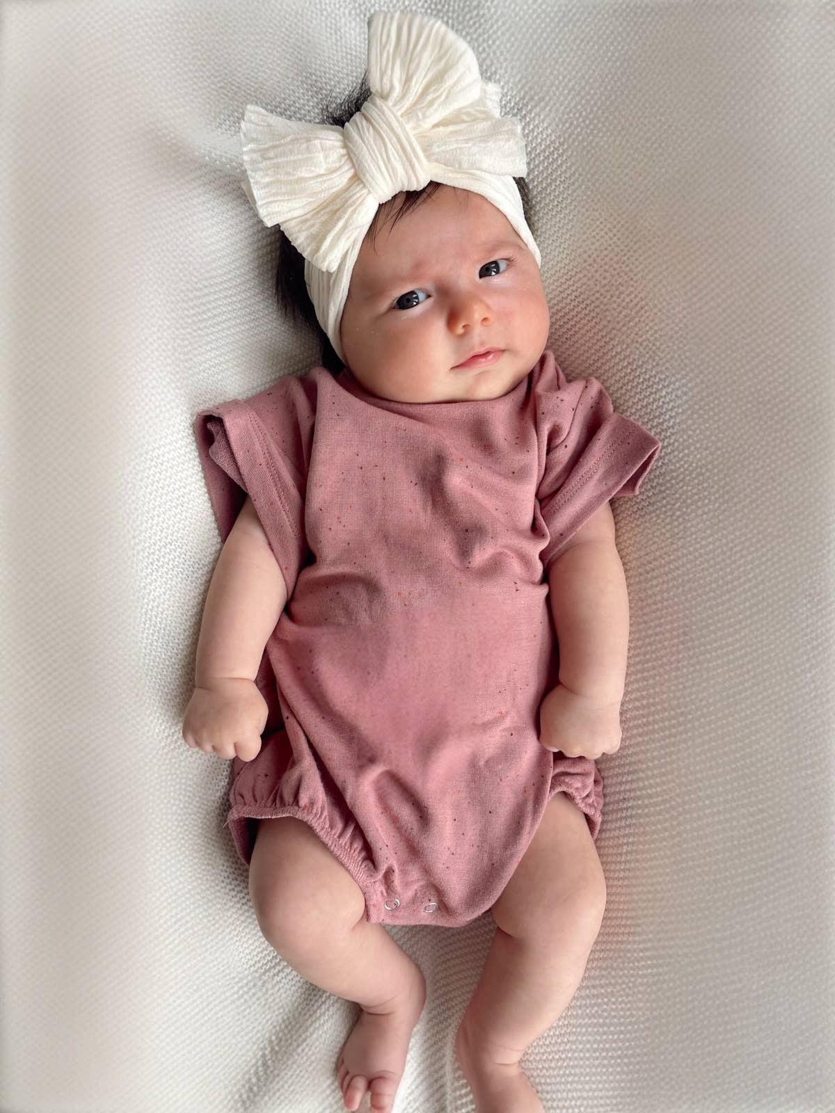 Baby in mauve speckle t-shirt bubble outfit with a large white headband, lying on a soft blanket.