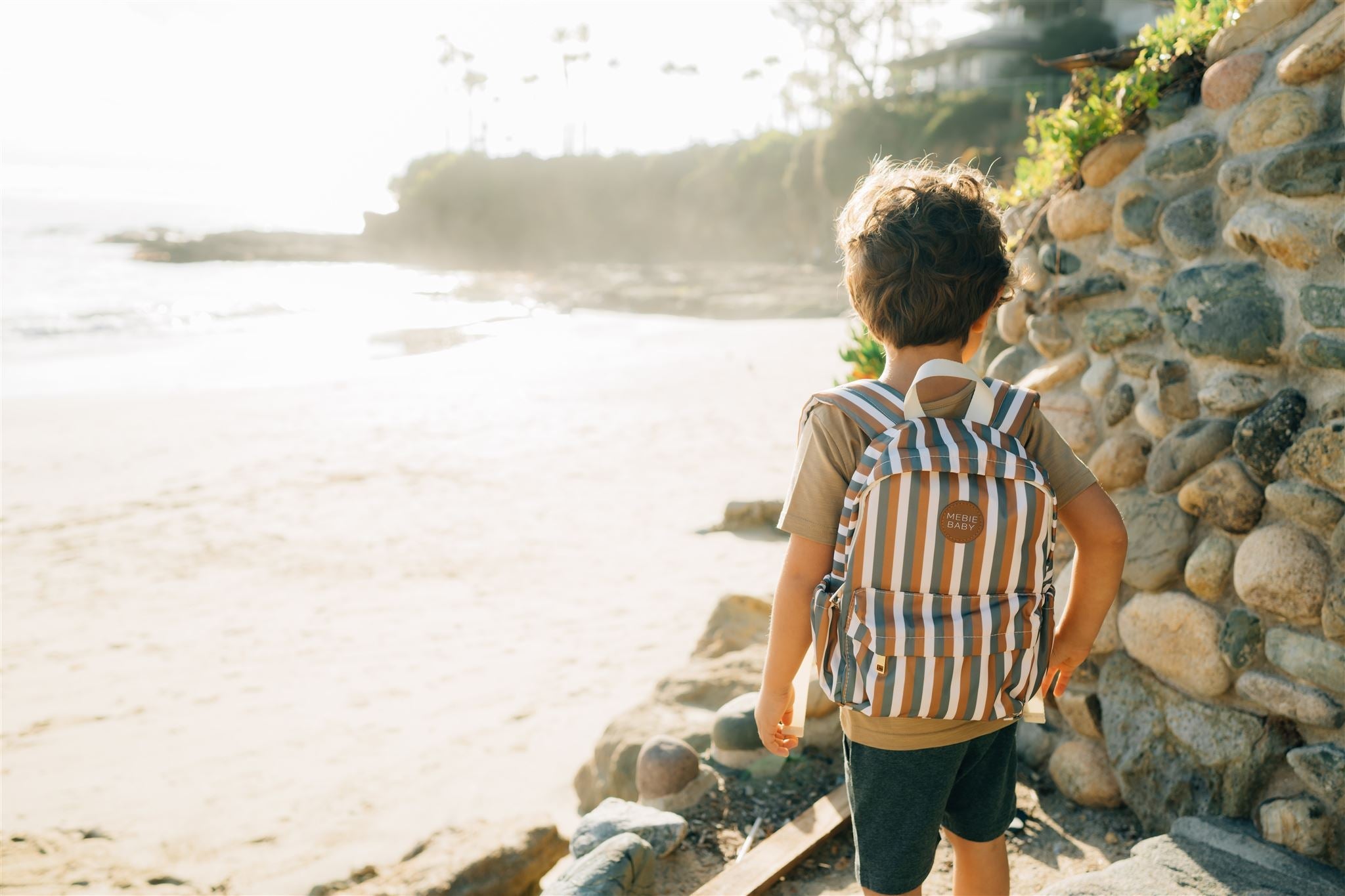 Child wearing a colorful sunset stripes mini backpack at the beach, perfect for kids' adventures.