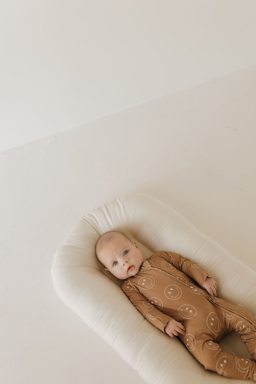 Baby wearing sandstone bamboo zip pajamas with smiley faces, resting comfortably on a soft surface.
