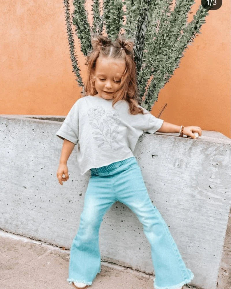 Toddler girl wearing light wash bell bottom jeans and grey tee, showcasing retro style in a trendy outdoor setting.