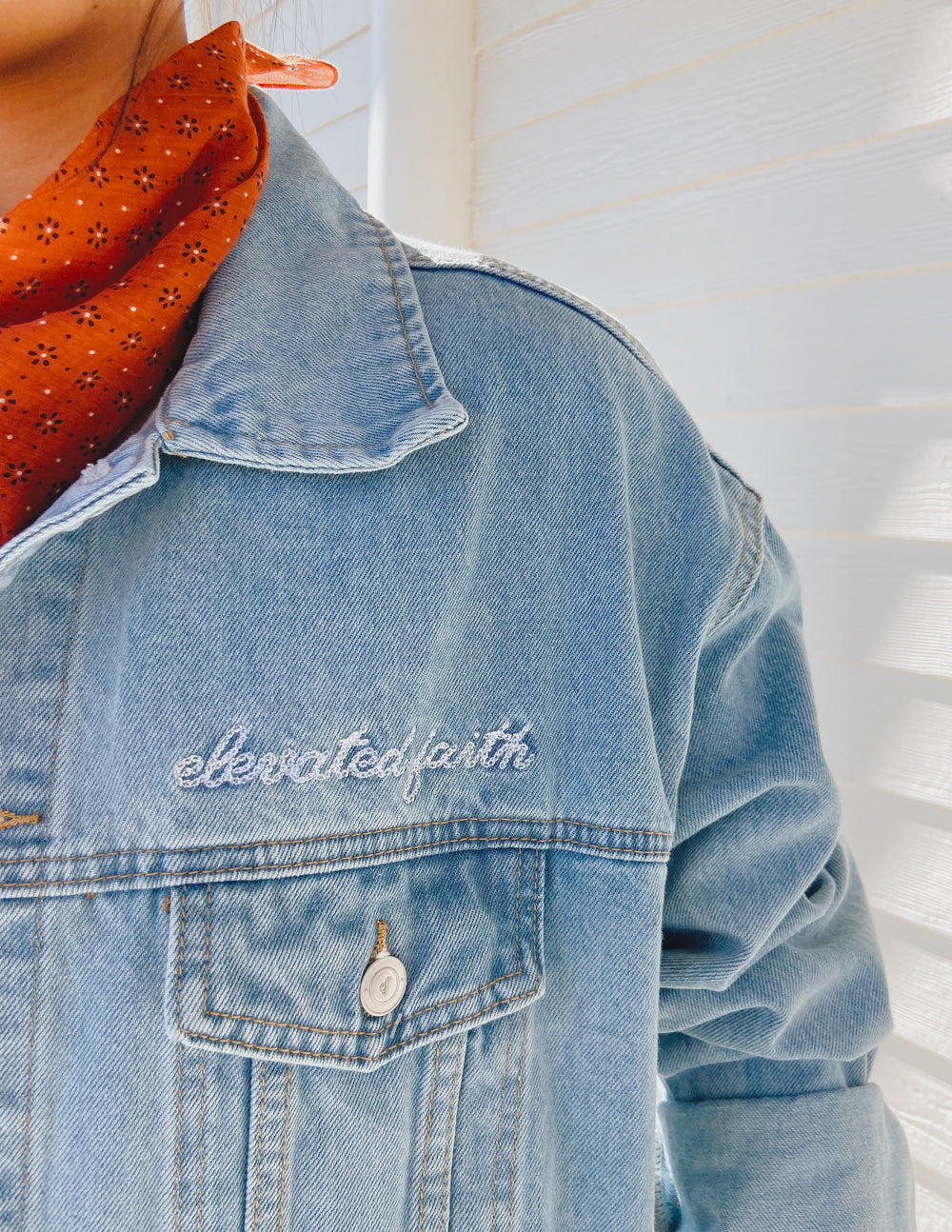 Close-up of women's denim jacket with 'elevatedfaith' embroidery, styled with a trendy scarf.