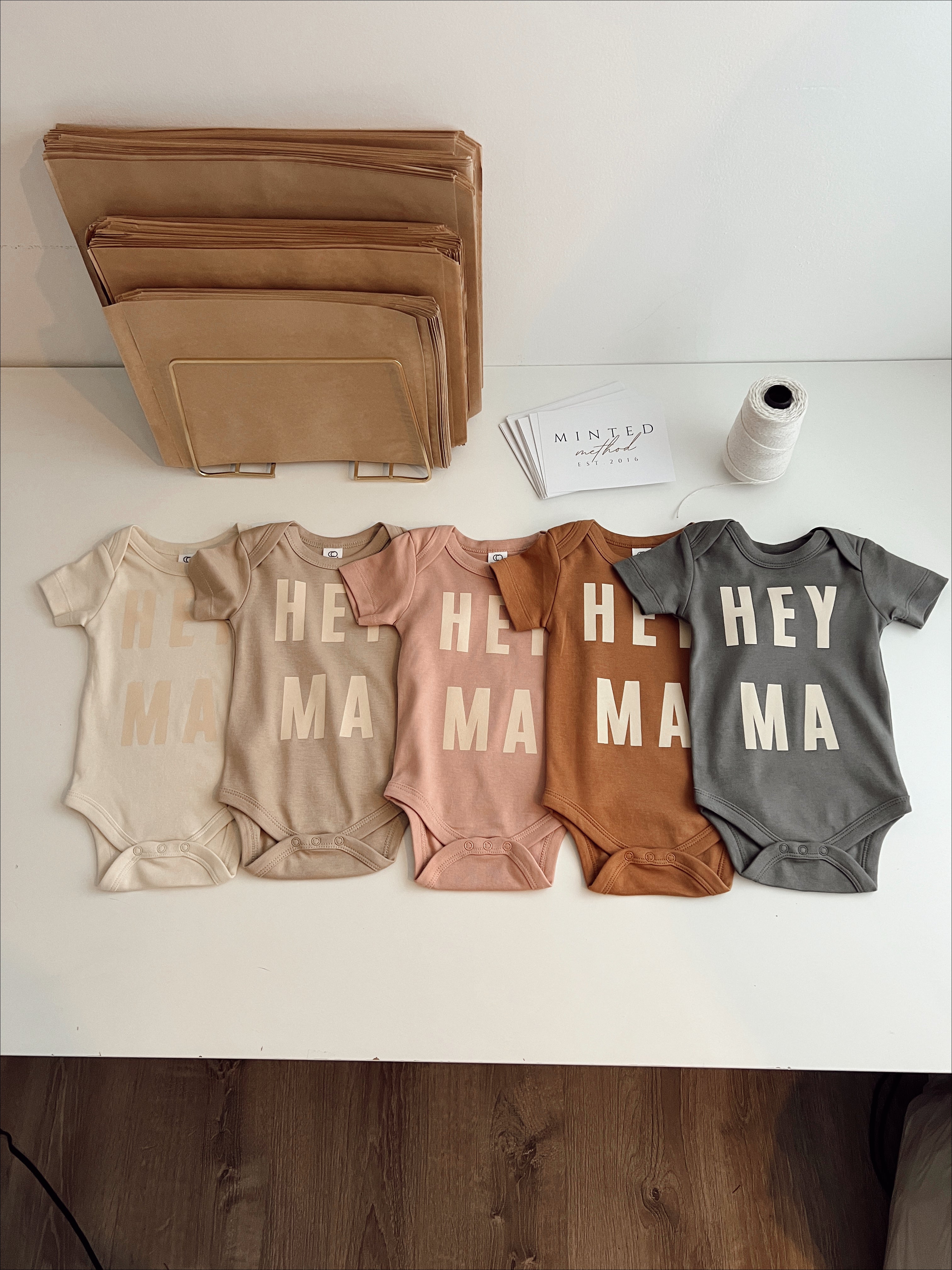 Display of Hey Ma organic baby onesies in cream, pink, brown, and gray, showcasing trendy and gender neutral baby clothes.