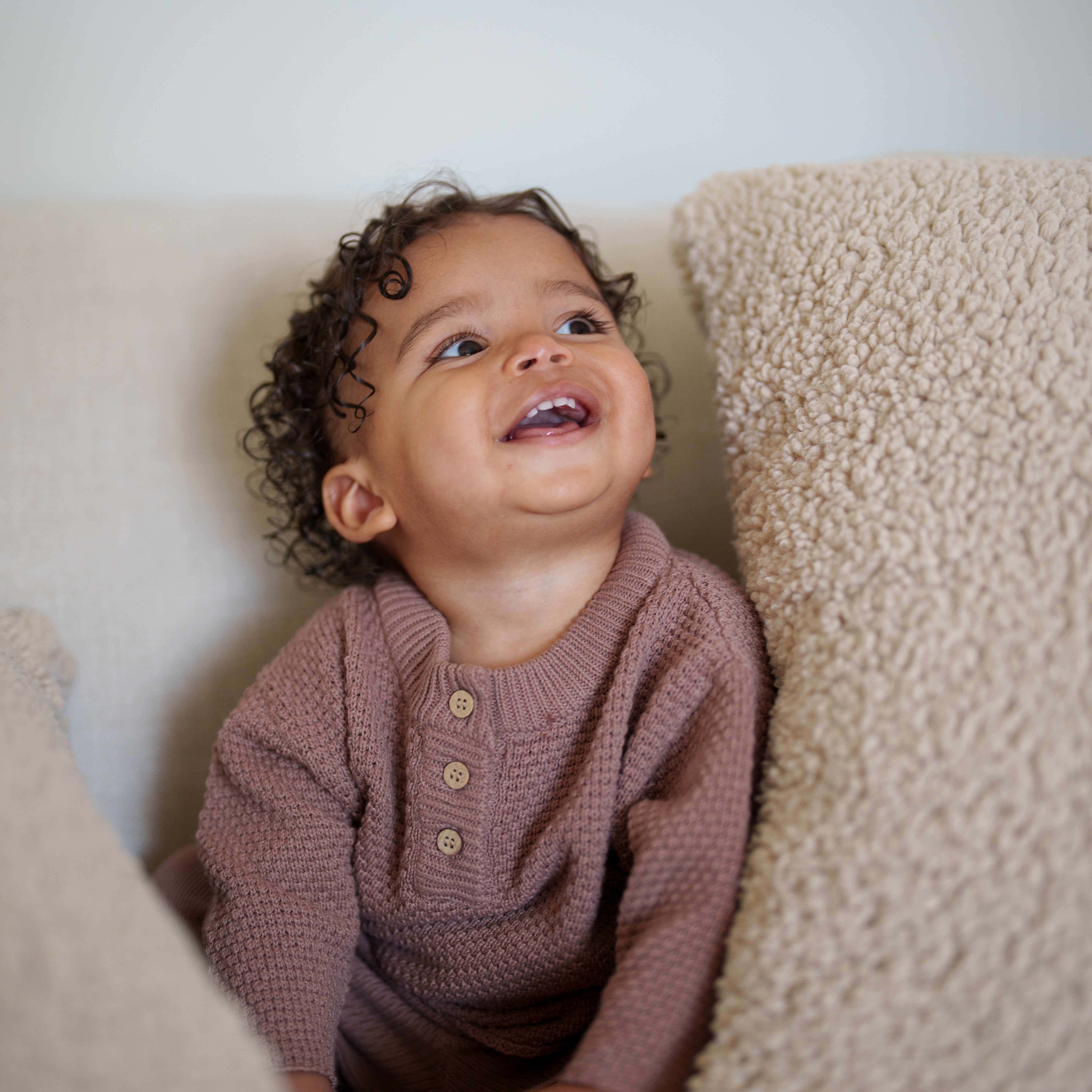 Smiling baby wearing Organic Knit Baggy Longsleeve in warm neutral tone sitting on a cozy couch.