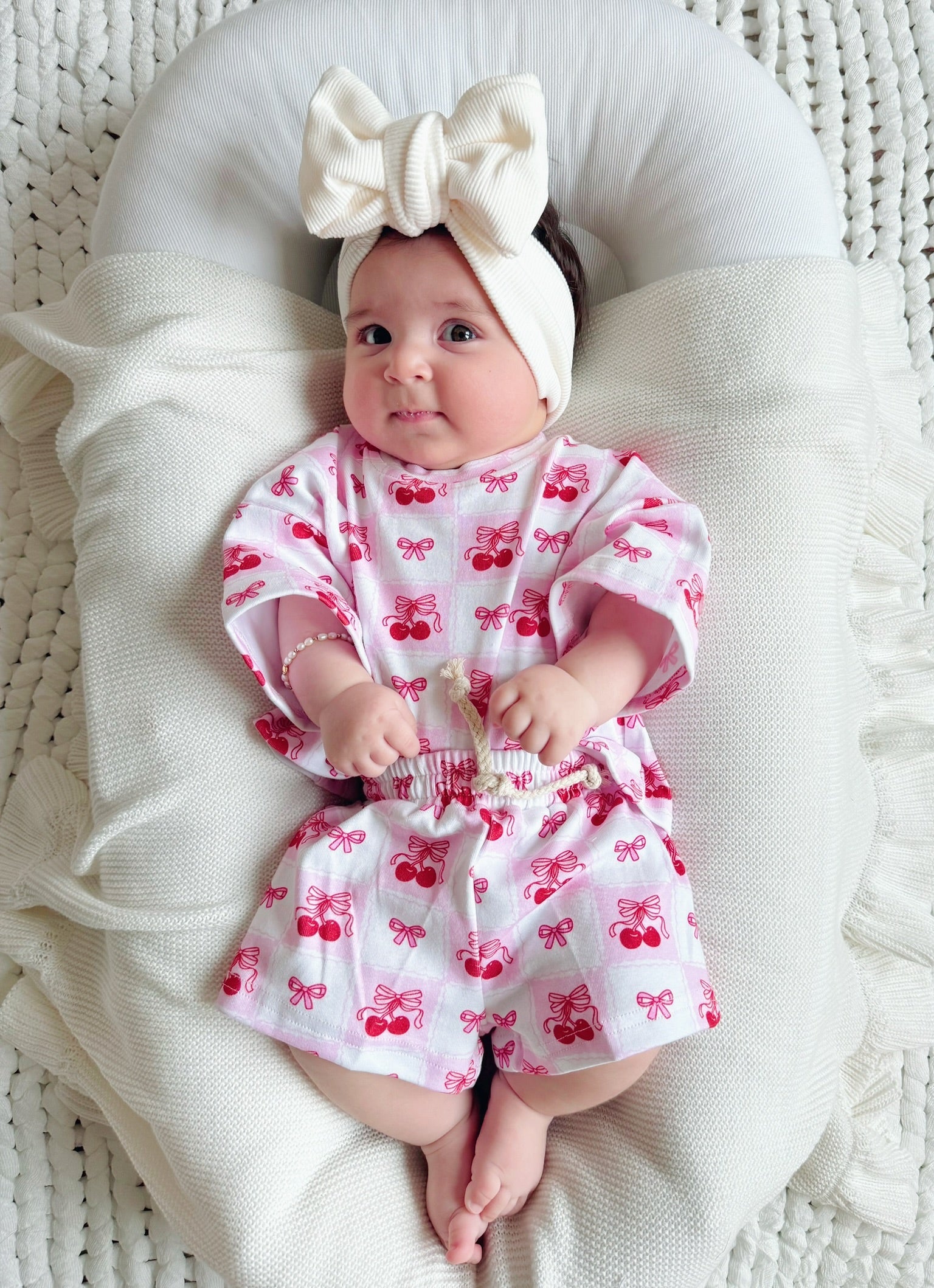 Baby wearing a checkered cherry shorts set with a headband, lying on a cozy blanket.