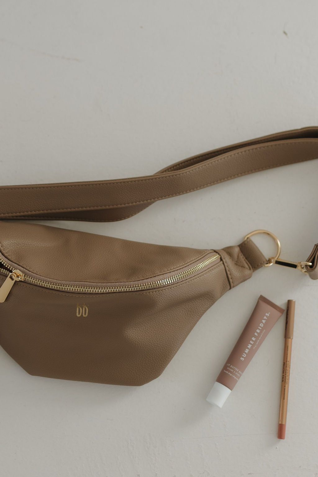 ff belt bag taupe laying flat with accessories, including lip products and a cosmetic pencil, showcasing its stylish design.