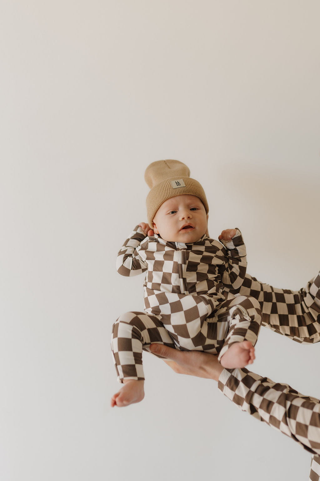 Baby wearing brown and white checkerboard bamboo zip pajamas by Dutton, held by a person.