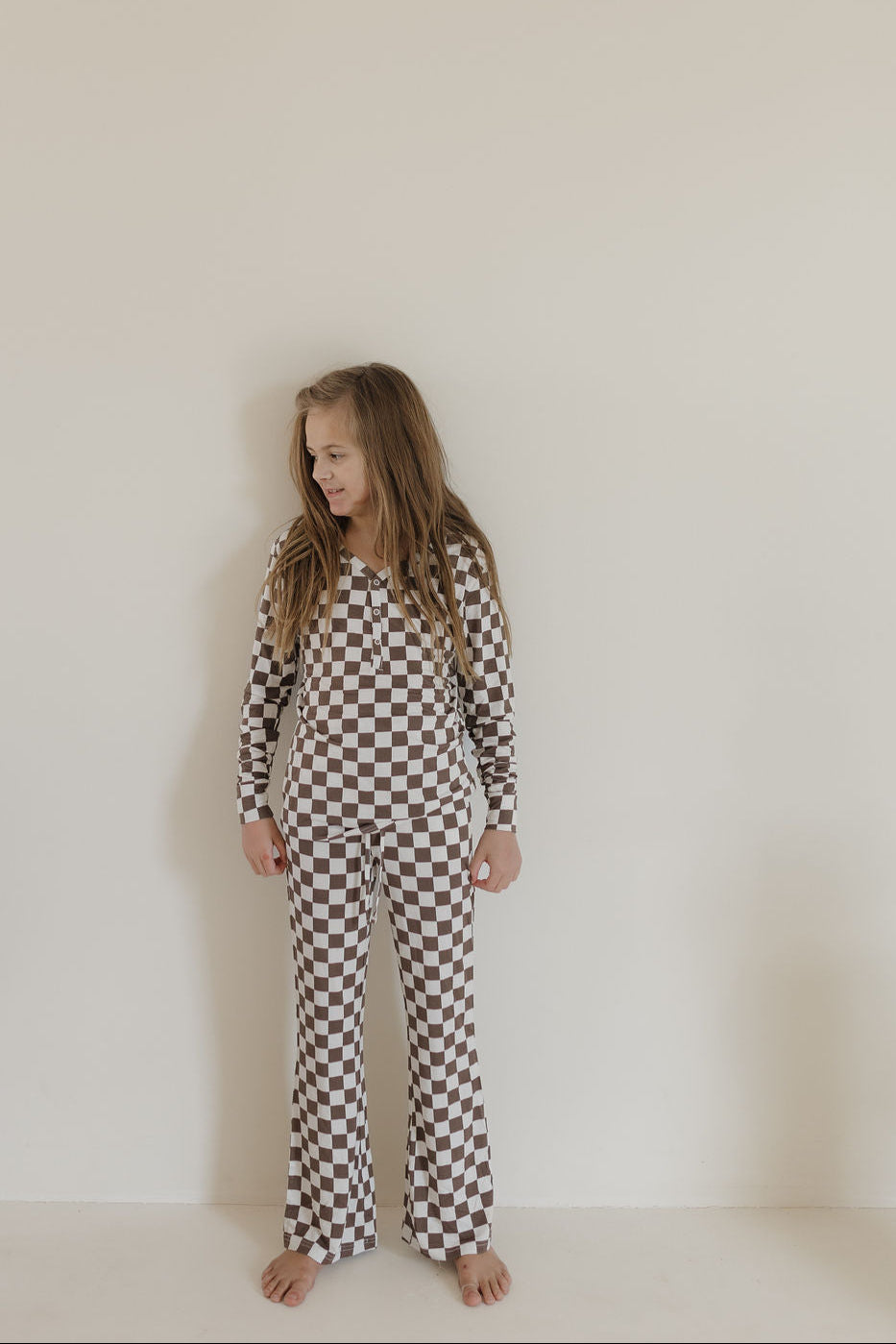 Girl wearing youth flare bamboo pajamas Dutton in brown-and-white checkerboard print, standing against a plain wall.