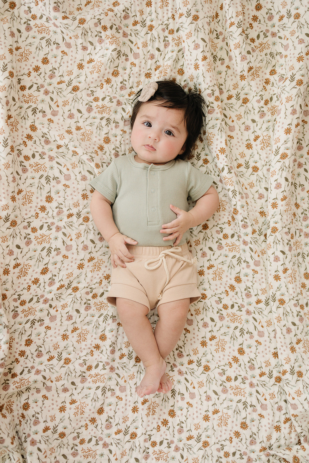 Infant wearing sage organic baby bodysuit and beige shorts, lying on floral-patterned blanket.