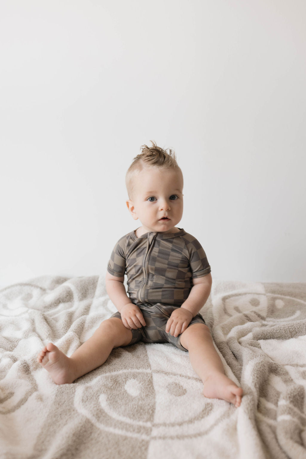 Baby in faded brown checkerboard bamboo zip shortie on a cozy blanket, showcasing trendy summer outfit.