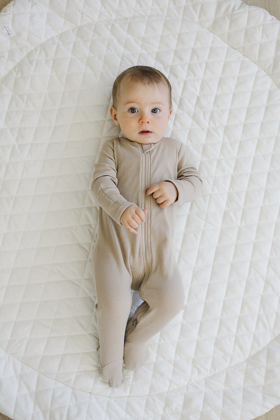 Baby in oatmeal organic cotton ribbed zipper pajamas on a soft quilted blanket.