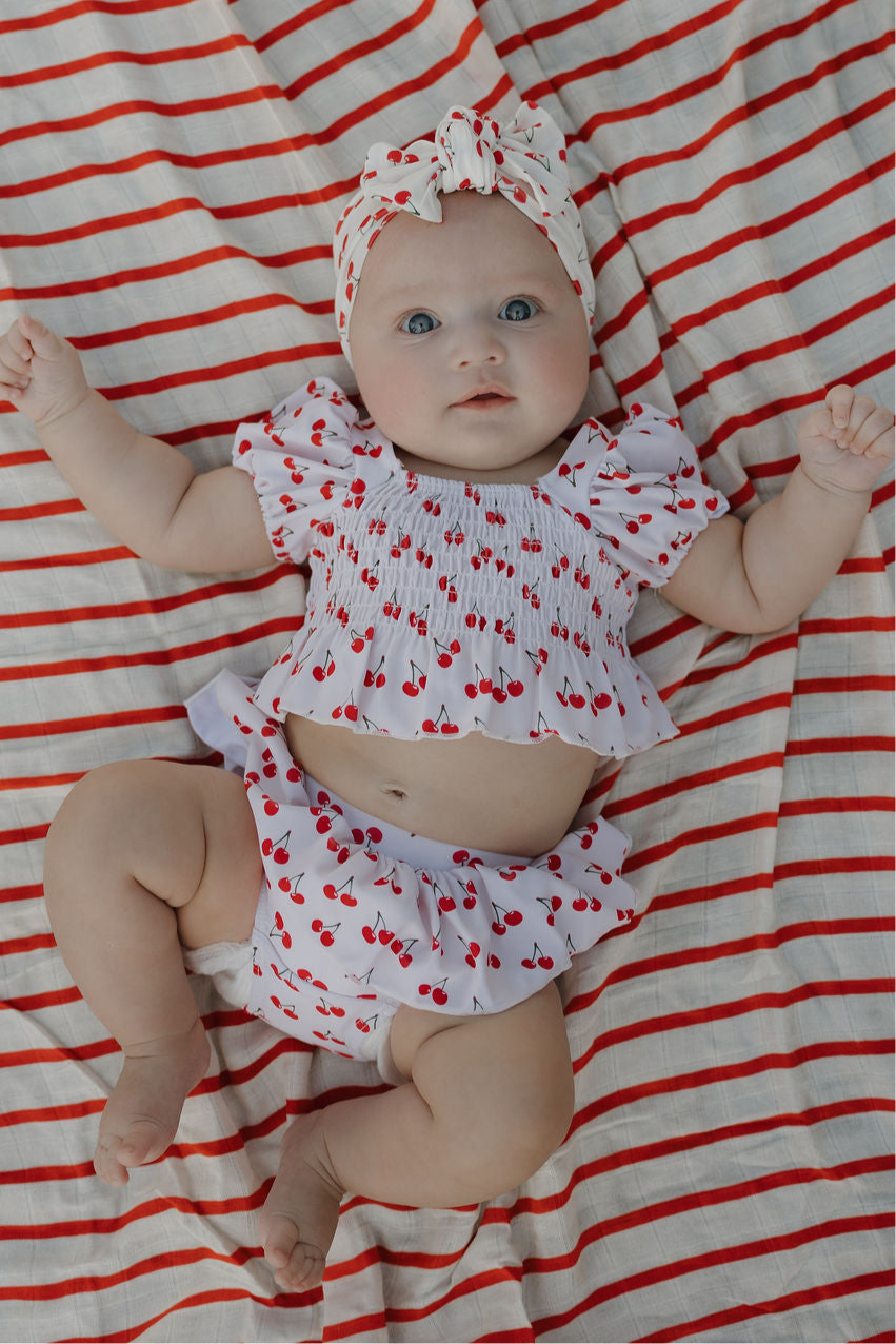 Baby wearing Sweetheart Cherries two-piece swimsuit with cherry print and matching headband, posed on a striped blanket.