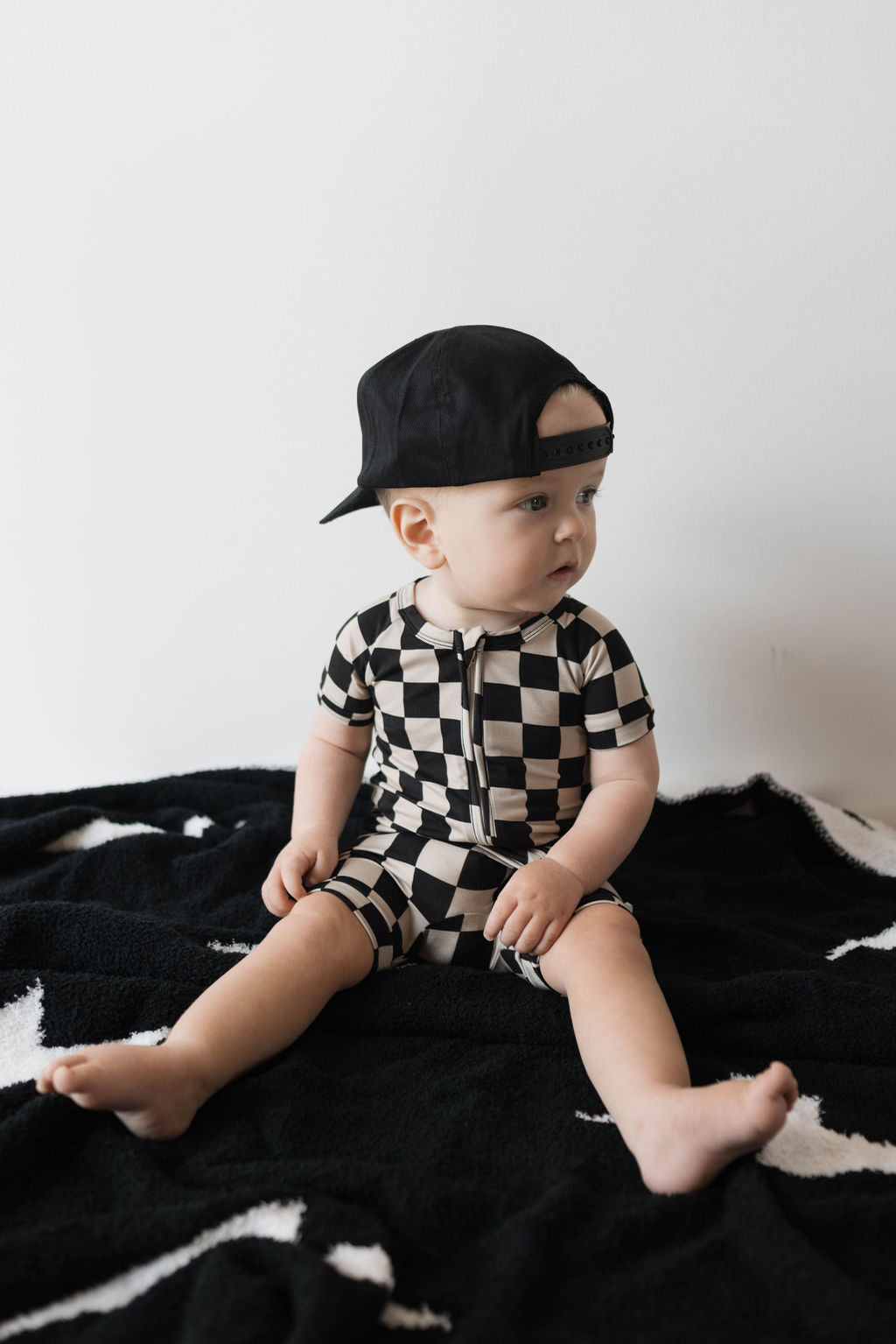 Baby wearing a black checkerboard bamboo zip shortie, sitting on a black blanket, stylish and trendy summer outfit.