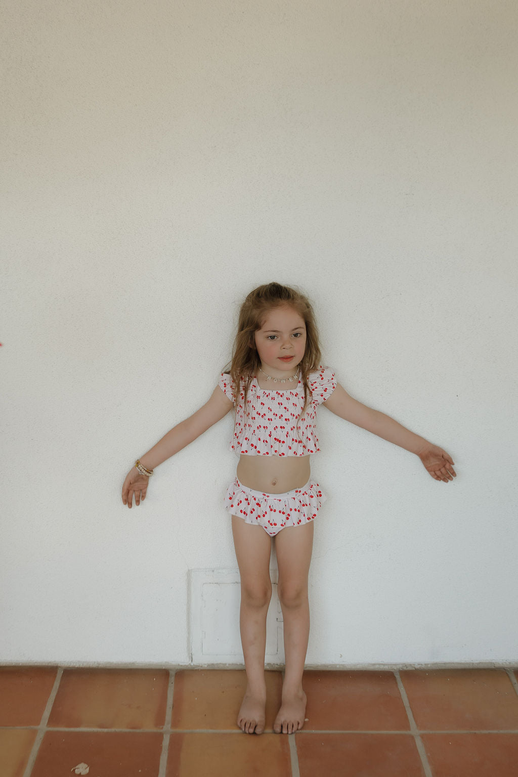 Cute girl in cherry print two-piece swimsuit with ruffle accents posing against a wall, perfect for summer beach fun.