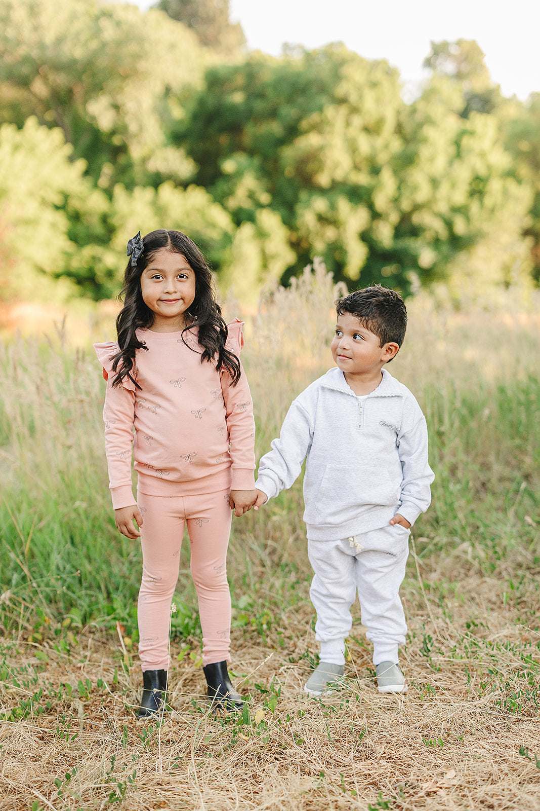Two children in a field, one wearing a pink ribbon ruffle set and the other in a white outfit, holding hands.