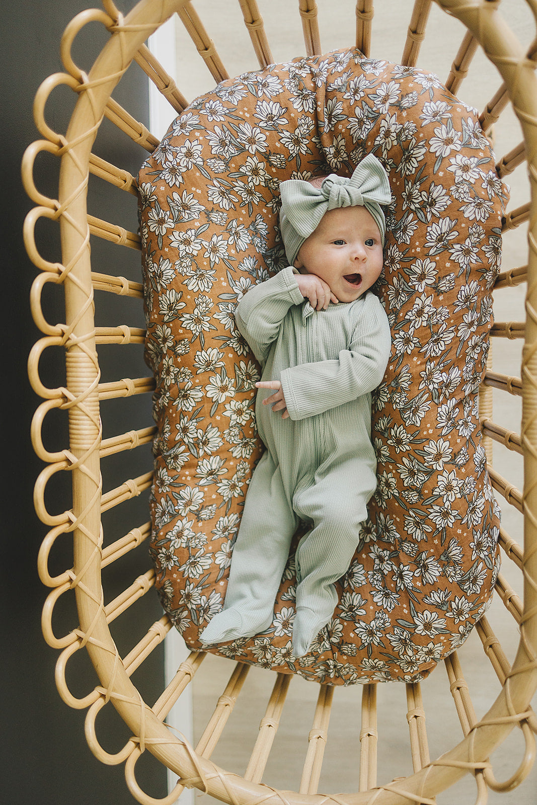 Baby in sage organic cotton ribbed pajamas with a bow, lying in a floral-patterned baby nest.