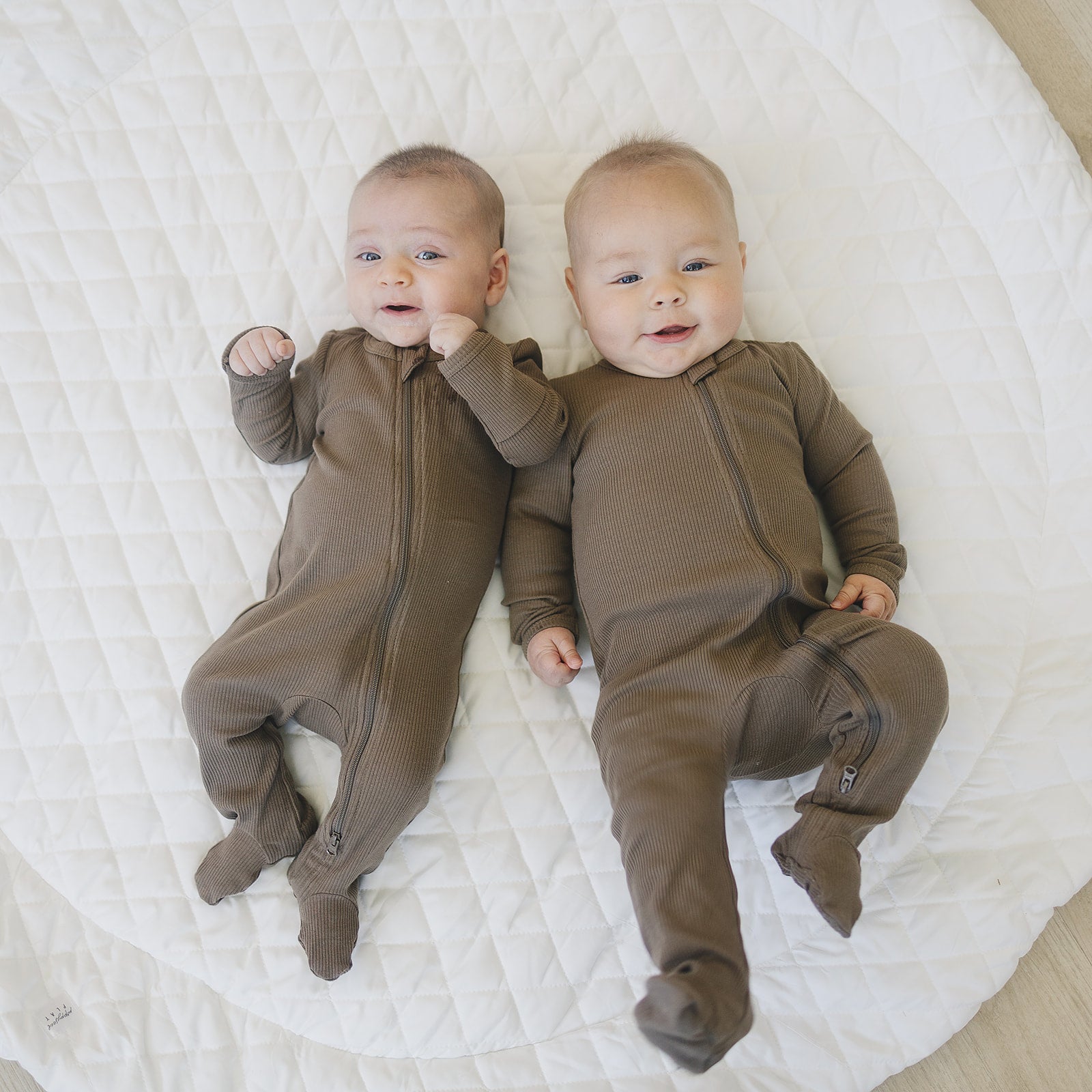 Two infants wearing cocoa organic baby pajamas with zip-up design on a quilted surface.