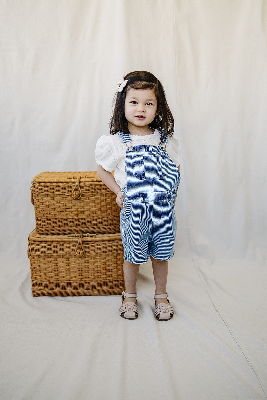 Toddler girl in blue striped short overalls, styled with a white top and cute accessories, perfect summer baby outfit.